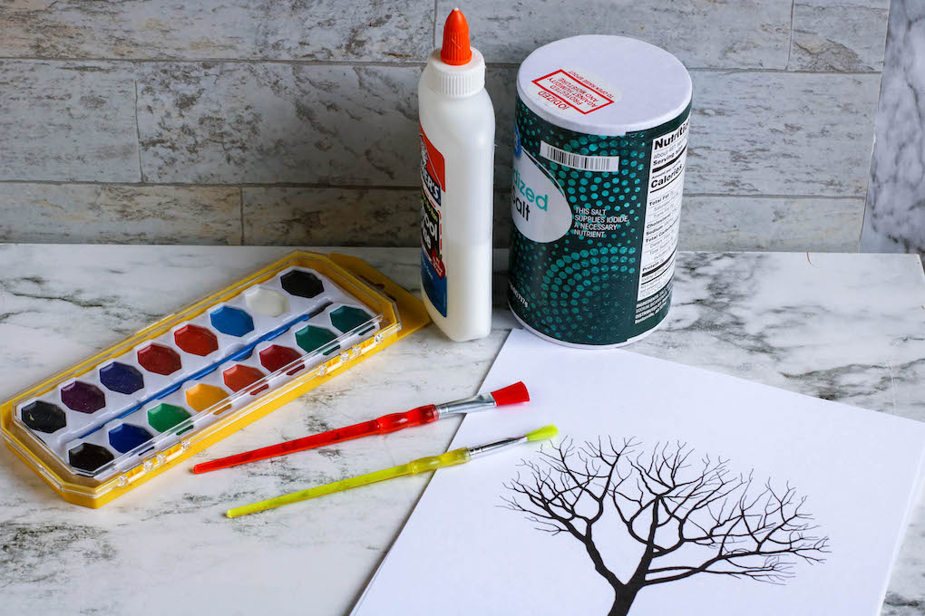 Watercolor paint set, glue, paintbrushes, and a canister of salt next to a line art tree template 