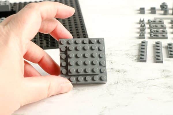 A hand holding a square grey LEGO piece and other grey LEGO pieces in the background