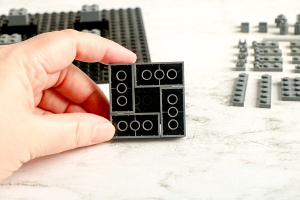 A hand holding a structure made of black LEGO bricks with bricks and a baseplate in the background