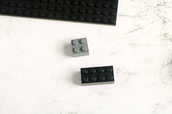 Two LEGO bricks, one grey and one black, near a corner of a black baseplate