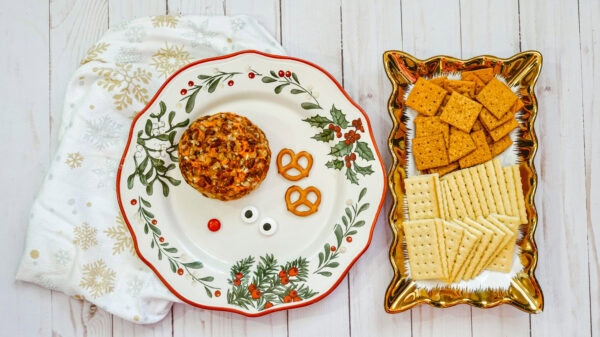 A holiday-themed plate holds the ingredients to assemble a Rudolph cheese ball, including a round cheese ball, two pretzels, candy eyes, and a red candy nose. A tray next to the plate is filled with square and rectangular crackers, ready for serving.