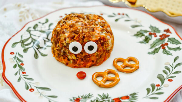 A partially assembled Rudolph cheeseball sits on a festive holiday plate. The cheese ball has candy eyes attached, with a red candy nose and two pretzels for antlers placed in front, ready to be added.