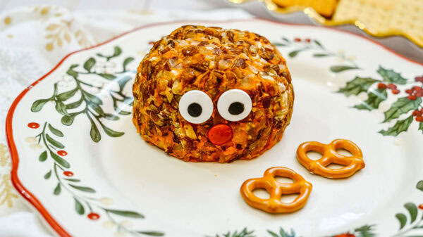 A Rudolph cheeseball with candy eyes and a red candy nose is partially assembled on a festive holiday plate. Two pretzels for antlers are placed in front of the cheeseball, ready to be added.