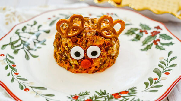 A fully assembled Rudolph cheeseball sits on a festive holiday plate. The cheeseball is decorated with candy eyes, a red candy nose, and pretzel antlers, ready for serving.