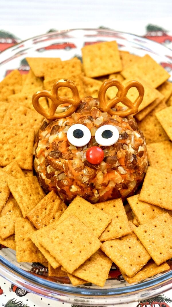 A festive cheese ball decorated to look like Rudolph, featuring pretzel antlers, candy eyes, and a red candy nose, surrounded by square crackers on a serving plate.