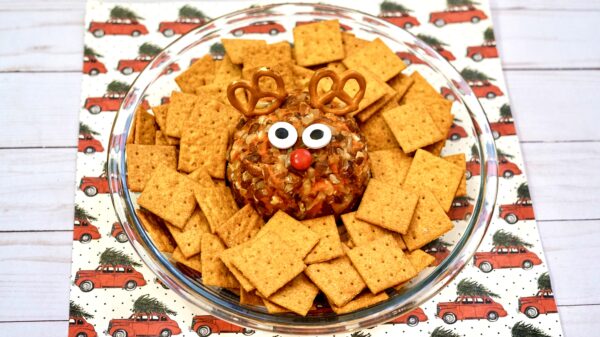 A festive Rudolph cheeseball is surrounded by square crackers on a clear serving platter. The cheeseball is decorated with pretzel antlers, candy eyes, and a red candy nose, placed on a holiday-themed background with red trucks carrying Christmas trees.
