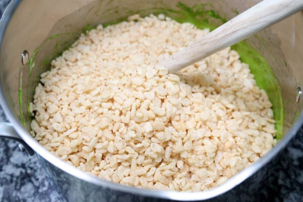 Pot filled with rice cereal being mixed into a green melted mixture using a wooden spoon