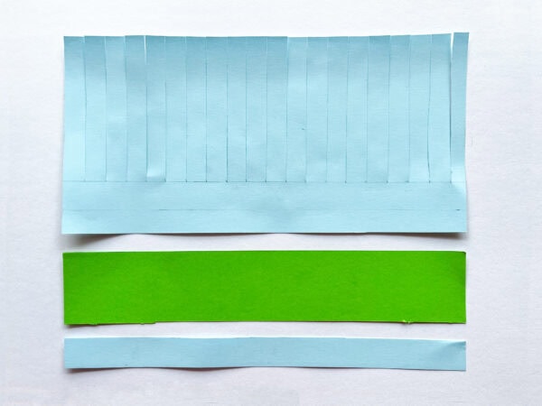 Strips of blue paper and a green rectangle are laid out, with the blue paper partially cut into vertical strips