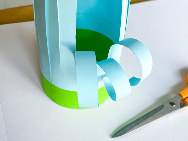 Blue strips are curled outward from the cylindrical paper basket using scissors