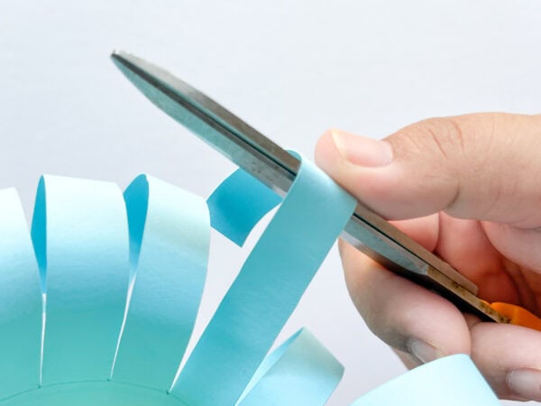 Blue strips are curled outward from the cylindrical paper basket using scissors