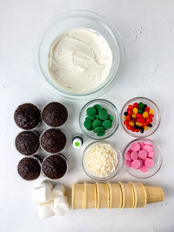 Ingredients for cupcake cones, including frosting, cupcakes, marshmallows, shredded coconut, jelly beans, pink and green candy melts, ice cream cones, and green food coloring.