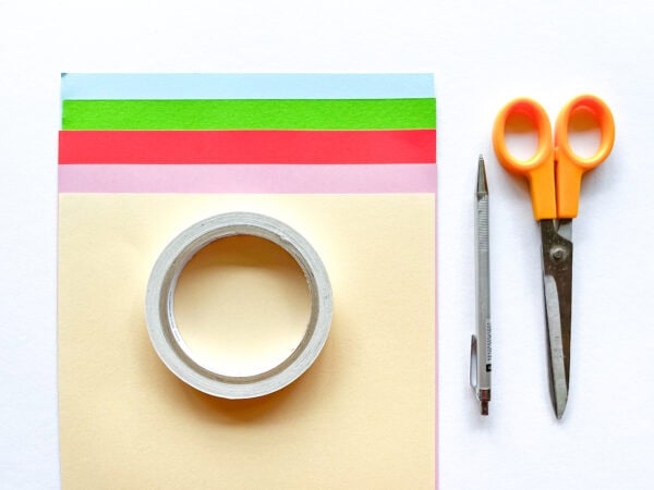 Craft materials laid out: colored paper sheets (blue, green, red, pink, beige), double-sided tape, scissors, and a pen