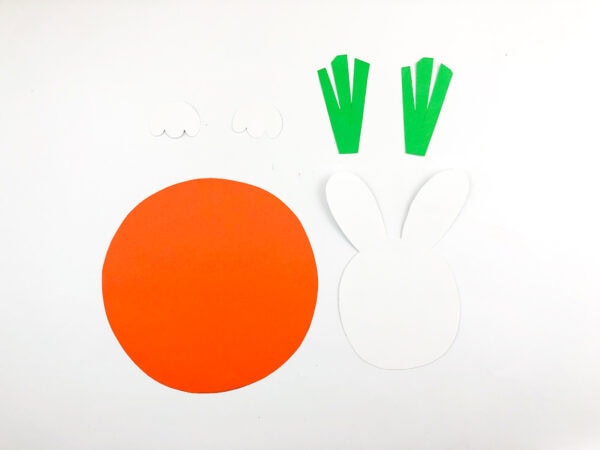 Cut-out paper pieces for a bunny-in-a-carrot craft, including a round orange piece, white bunny face, green carrot tops, and white paws.