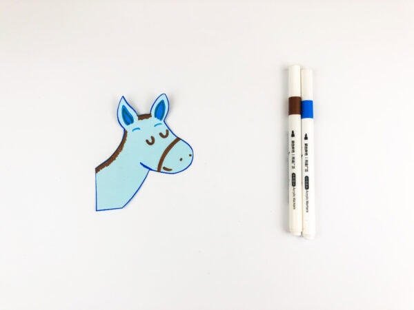A cutout of a donkey's head drawn on light blue paper with brown and blue marker details, next to two markers.