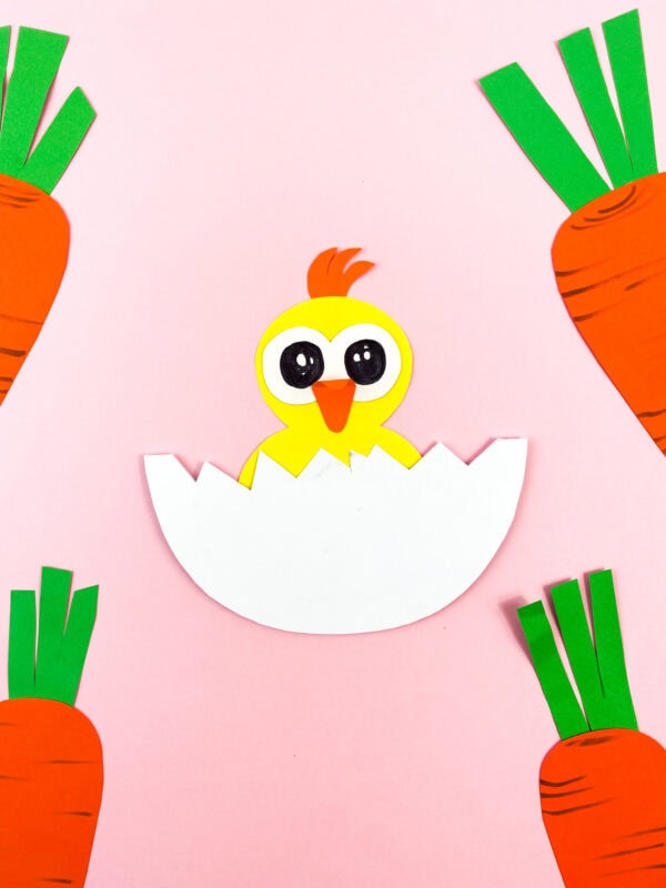 The completed chick craft surrounded by paper carrots on a pink background.