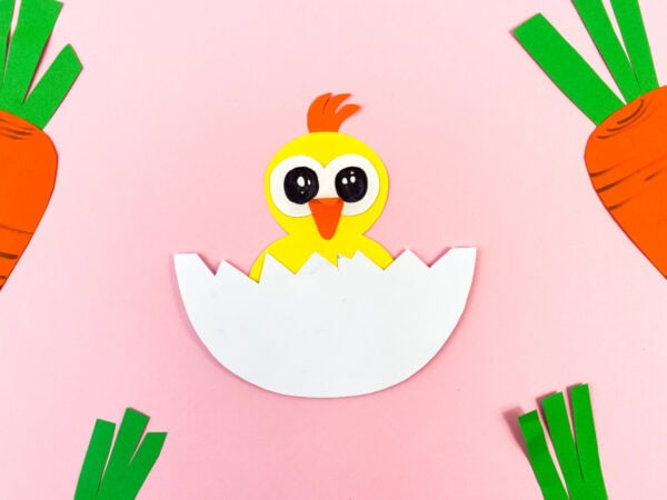 A paper chick hatching from an egg with paper carrots on a pink background.