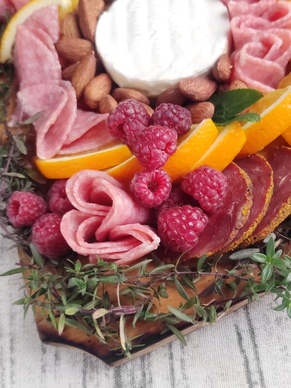 A close-up of the charcuterie board corner, highlighting salami roses, raspberries, almonds, and orange slices.