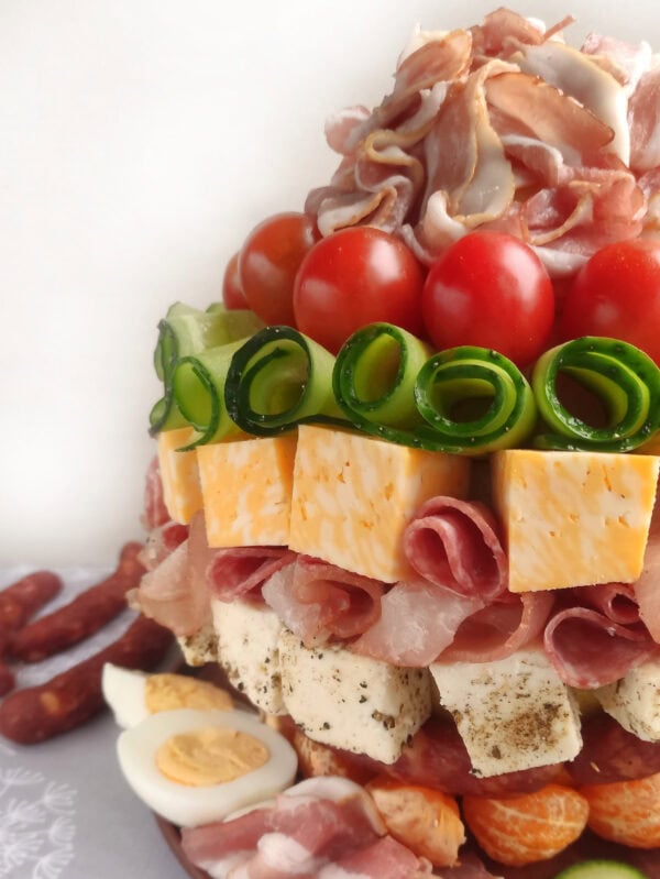 Close-up view of a charcuterie tower featuring rolled meats, cherry tomatoes, cucumber spirals, marbled cheese cubes, and diced young cheese with herbs