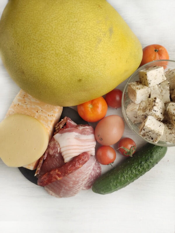 Assortment of ingredients including a large pomelo, diced young cheese with herbs in a bowl, sliced cheeses, various cured meats, whole cherry tomatoes, eggs, a cucumber, and tangerines