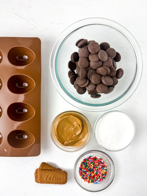 Ingredients for chocolate egg treats, including chocolate melts, peanut butter, powdered sugar, sprinkles, and a Lotus Biscoff cookie, arranged with a silicone egg mold.