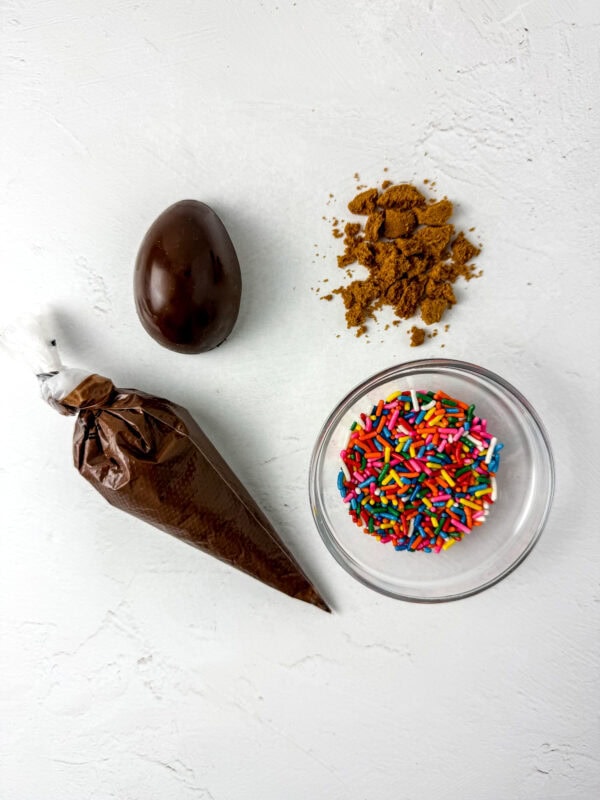 Ready for decorating! A smooth chocolate egg alongside crushed Biscoff cookies, colorful sprinkles, and a piping bag filled with melted chocolate for drizzling.