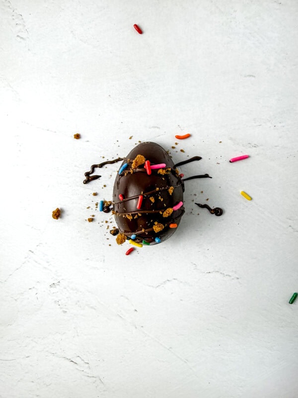 A chocolate egg drizzled with melted chocolate and topped with colorful sprinkles and crushed Biscoff cookies, creating a festive and indulgent treat.