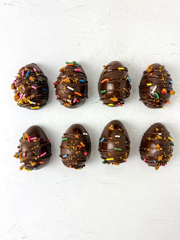 A batch of homemade chocolate eggs, drizzled with melted chocolate and sprinkled with colorful decorations and crushed Biscoff cookies, arranged neatly on a white surface.