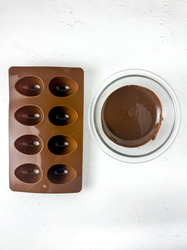 Silicone egg mold next to a glass bowl of melted chocolate on a white surface.