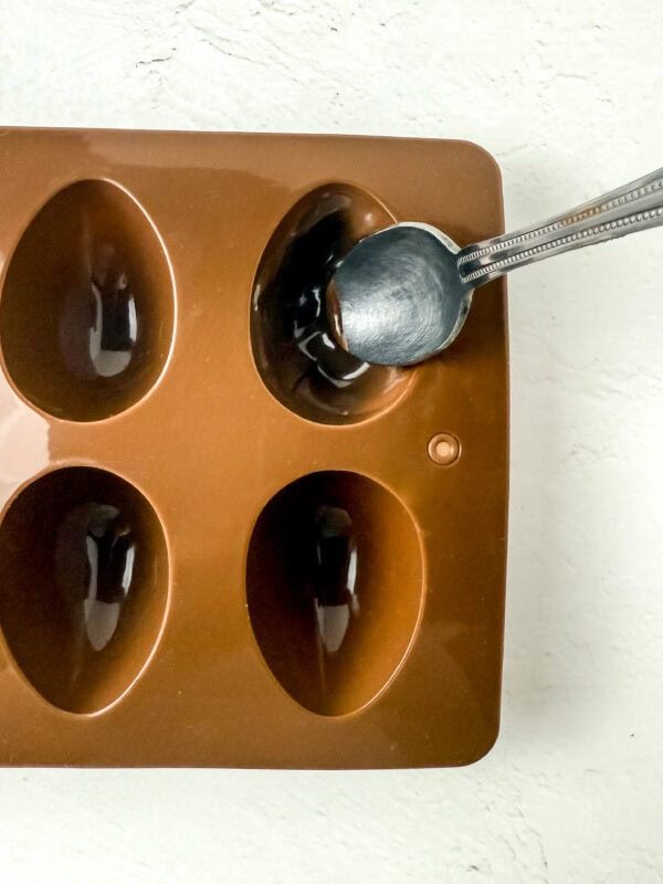 Close-up of a spoon spreading melted chocolate inside a silicone egg mold.