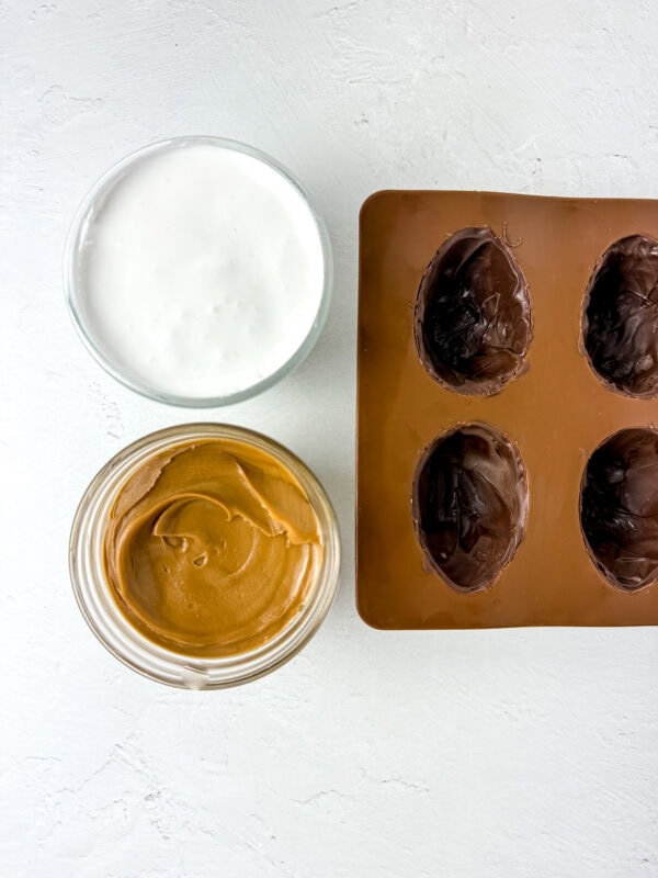 Jars of marshmallow fluff and cookie butter next to a chocolate-coated silicone mold, prepared for filling.