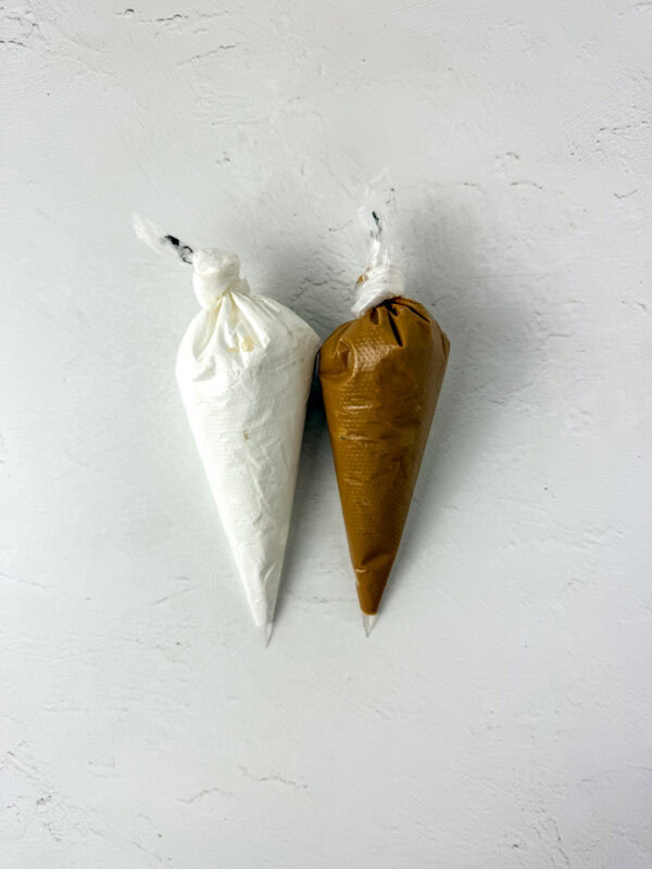 Two piping bags filled with marshmallow fluff (white) and cookie butter (brown), tied at the top and placed on a white surface.