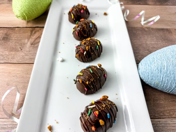 Five chocolate-covered eggs drizzled with chocolate and topped with crushed Biscoff cookies and colorful sprinkles, arranged on a white rectangular plate with decorative pastel Easter eggs and ribbons in the background.