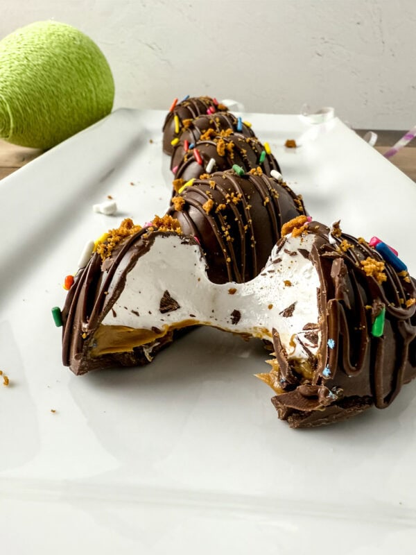 A close-up of a chocolate egg with a bite taken out, revealing layers of fluffy marshmallow and creamy cookie butter inside. The chocolate shell is drizzled with more chocolate and sprinkled with colorful decorations and crushed Biscoff cookies.