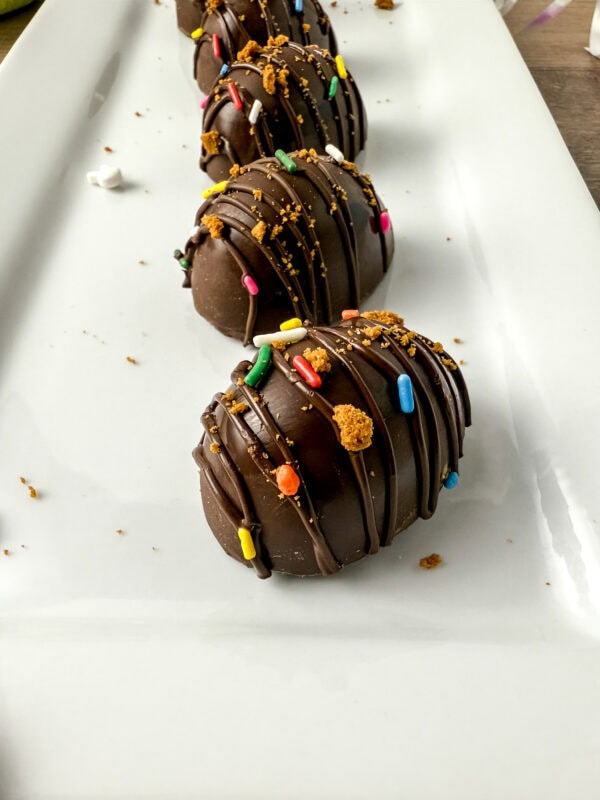 Close-up of chocolate-covered eggs drizzled with chocolate and topped with colorful sprinkles and crushed Biscoff cookies, arranged on a white rectangular plate.