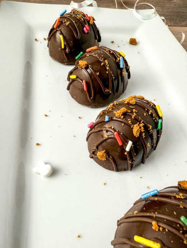 Chocolate-covered Easter eggs drizzled with chocolate and topped with colorful sprinkles and crushed Biscoff cookies, arranged on a white plate with scattered crumbs and marshmallow bits.
