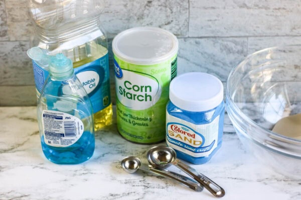 A collection of ingredients, including cornstarch, colored sand, dish soap, vegetable oil, and measuring spoons, placed on a countertop.