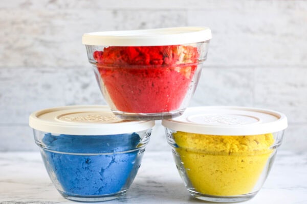 Three small glass containers with lids, each filled with kinetic sand in red, blue, and yellow.