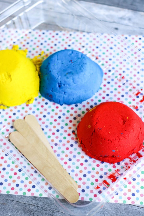 A closer look at the blue sand being cut with a wooden stick, with red sand nearby on a polka-dotted surface.