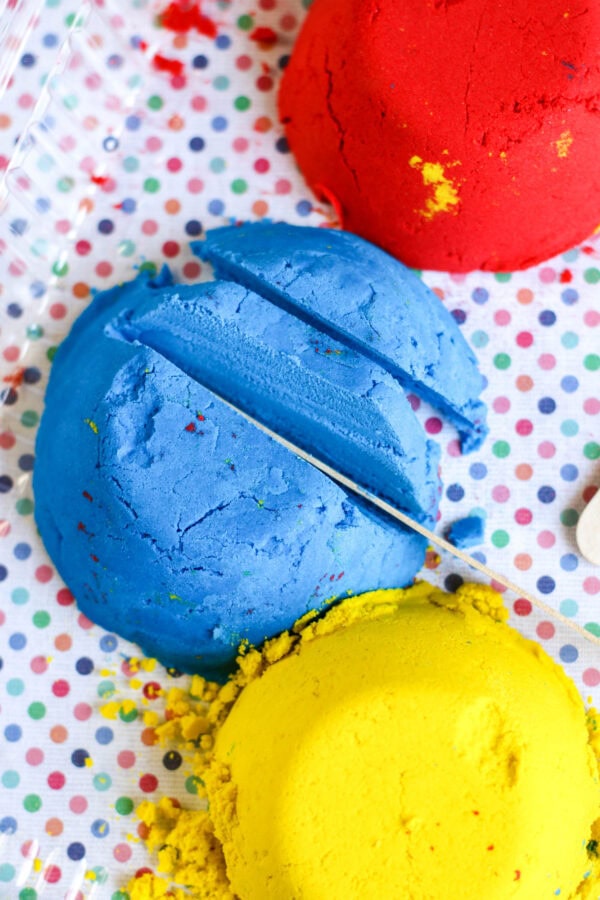 blue sand being cut with a wooden stick, with red sand nearby on a polka-dotted surface.