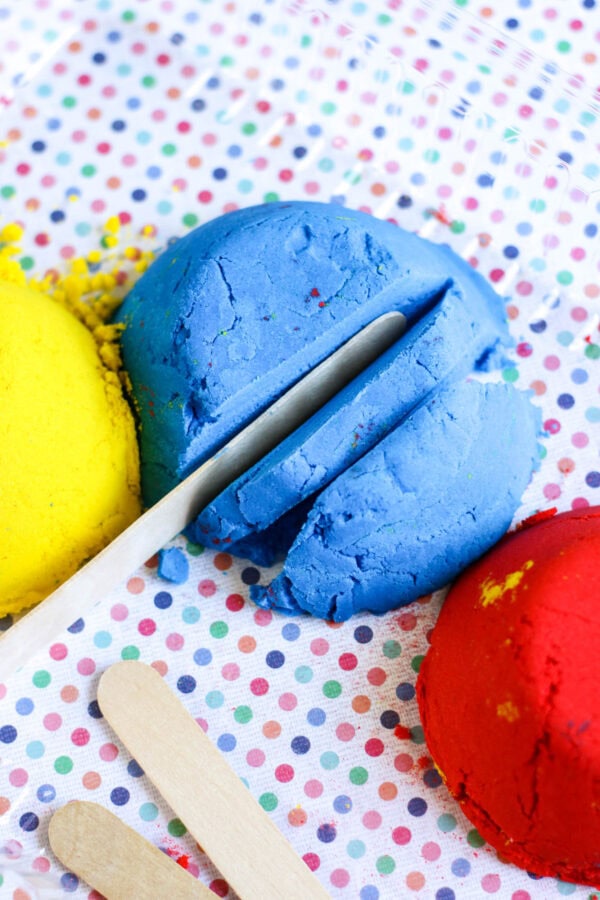 A closer look at the blue sand being cut with a wooden stick, with red sand nearby on a polka-dotted surface.