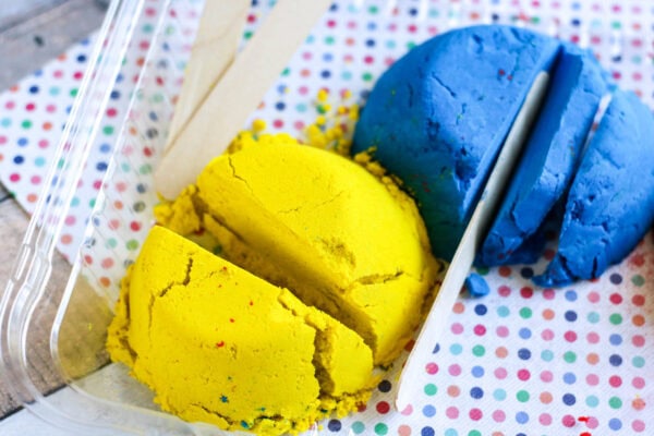 A close-up of blue and yellow kinetic sand being sliced with a popsicle stick
