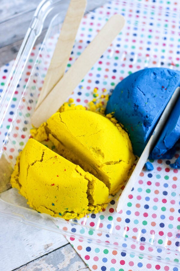 Yellow and blue kinetic sand being cut in a plastic container with wooden sticks beside them.