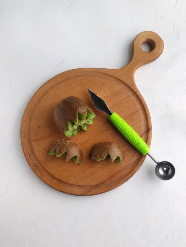 Kiwi halves cut with zigzag edges on a wooden board with a carving tool.