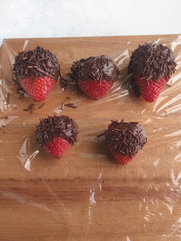 Strawberries dipped in chocolate and sprinkles on a wooden board with plastic wrap.