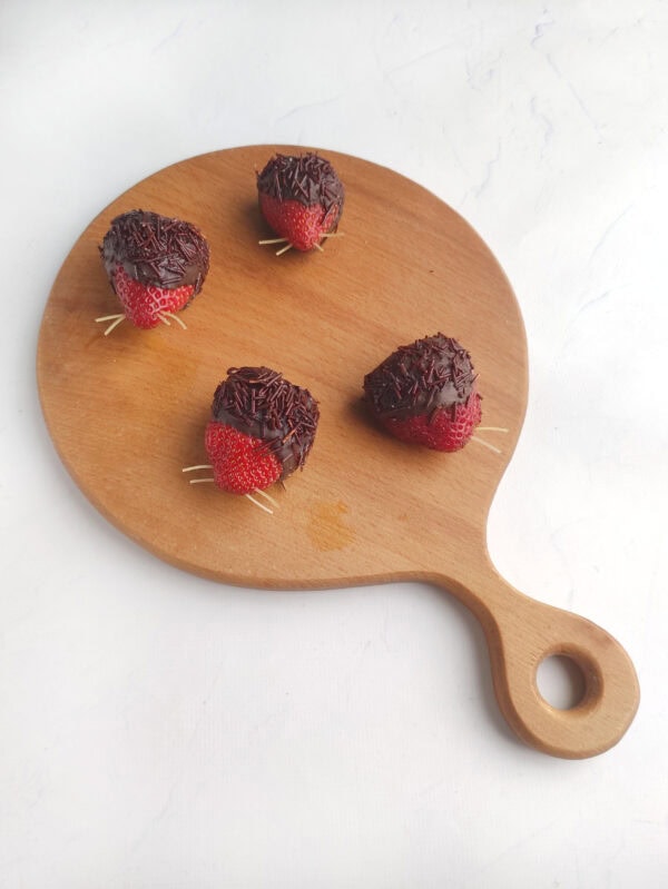 Chocolate-dipped strawberries with pasta whiskers placed on a round wooden board.