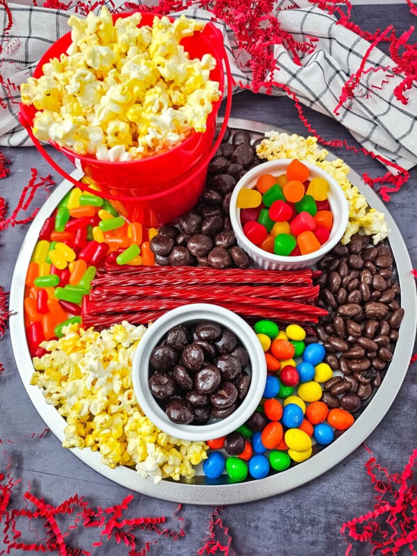 A circular snack tray filled with colorful candy, popcorn, licorice, and chocolates, featuring a red bucket of popcorn in the center.