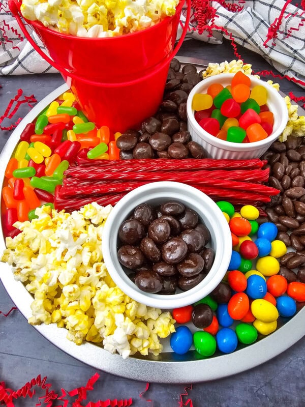 A close-up shot of the snack tray showing chocolate raisins, M&Ms, licorice, and gummy candies beside a popcorn bucket.