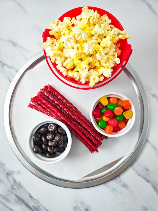 Popcorn, licorice, chocolates, and gummies arranged around the red popcorn bucket on a snack tray.