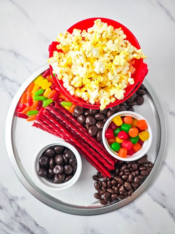 Snack board partially filled with licorice, chocolate raisins, popcorn, and colorful candies next to a popcorn bucket.
