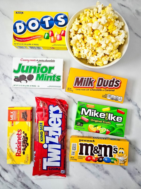 Boxes of various movie theater candies arranged around a bowl of popcorn on a marble surface.